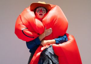 Woman dressed in clown makeup and a cowboy hat hugs a red inflatable sky dancer around her shoulders.
