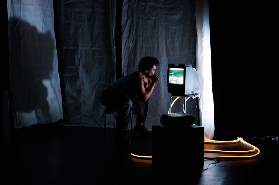 A man sits hunched forward on a chair in a dark stage space, intensely watching a glowing CRT television, surrounded by draped fabric, shadows, and looping cables on the floor.