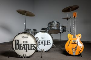 A drum kit branded with “The Beatles” on the bass drums stands in a minimalist gallery space beside a honey-colored Gretsch electric guitar, both displayed as museum-style music memorabilia against a neutral backdrop.