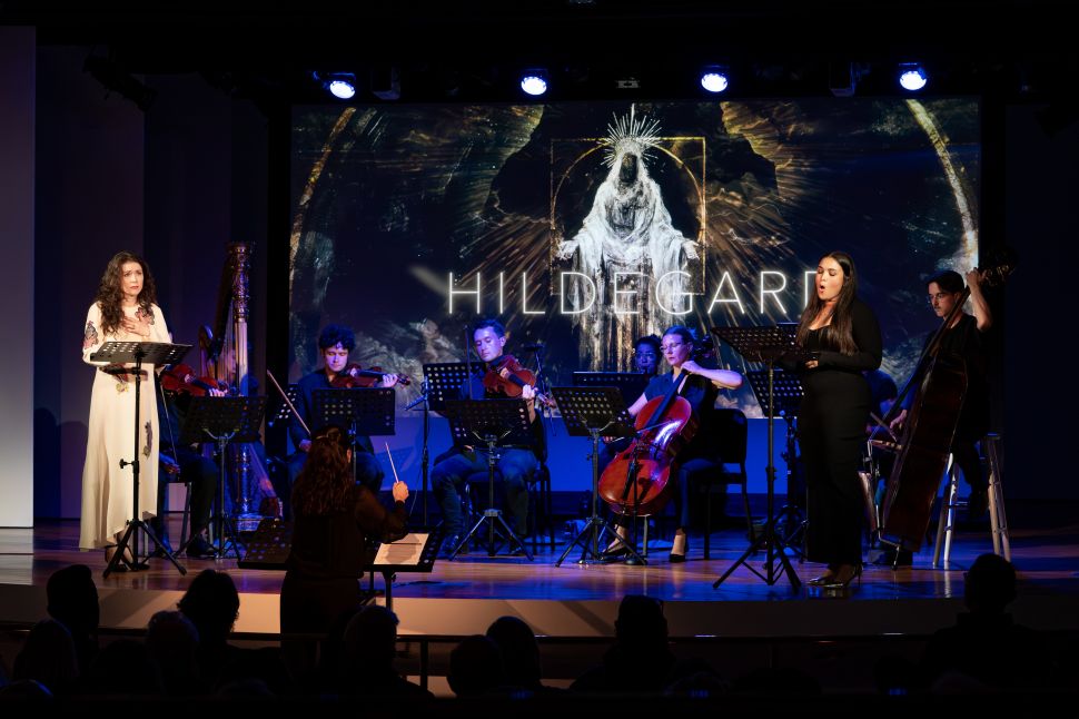 A live performance of Hildegard shows two vocalists and a chamber ensemble onstage, framed by a glowing digital projection of the opera’s title and imagery, capturing the work’s fusion of music, spirituality and contemporary visual design.
