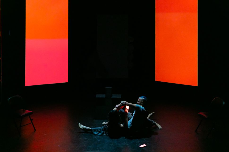 Two performers sit on a dark stage illuminated by large glowing red and orange screens, creating an abstract digital environment around them.