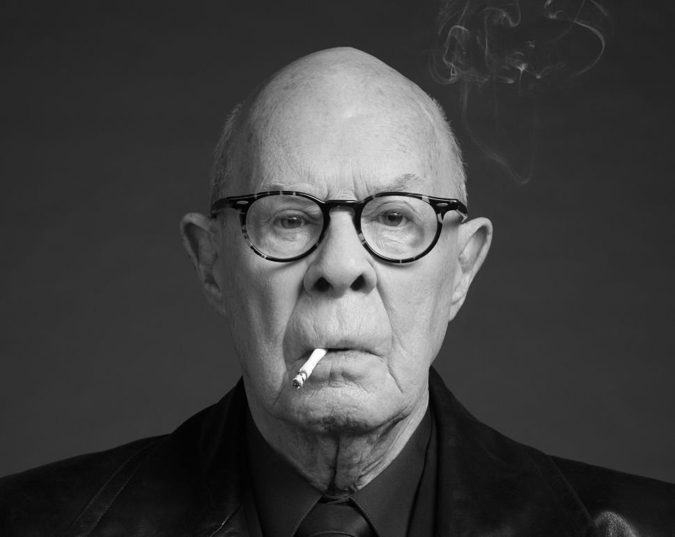 A black-and-white studio portrait shows an older man in glasses, a leather jacket and a tie looking straight at the camera with a lit cigarette between his lips, representing Hans van Manen.