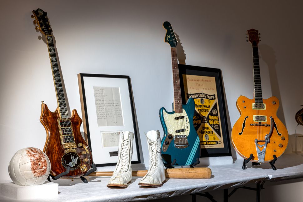 A tabletop display presents several iconic music and sports memorabilia items, including vintage electric guitars on stands, a framed handwritten document, a baseball bat, a worn white leather ball and a pair of white lace-up boxing boots arranged under gallery lighting.