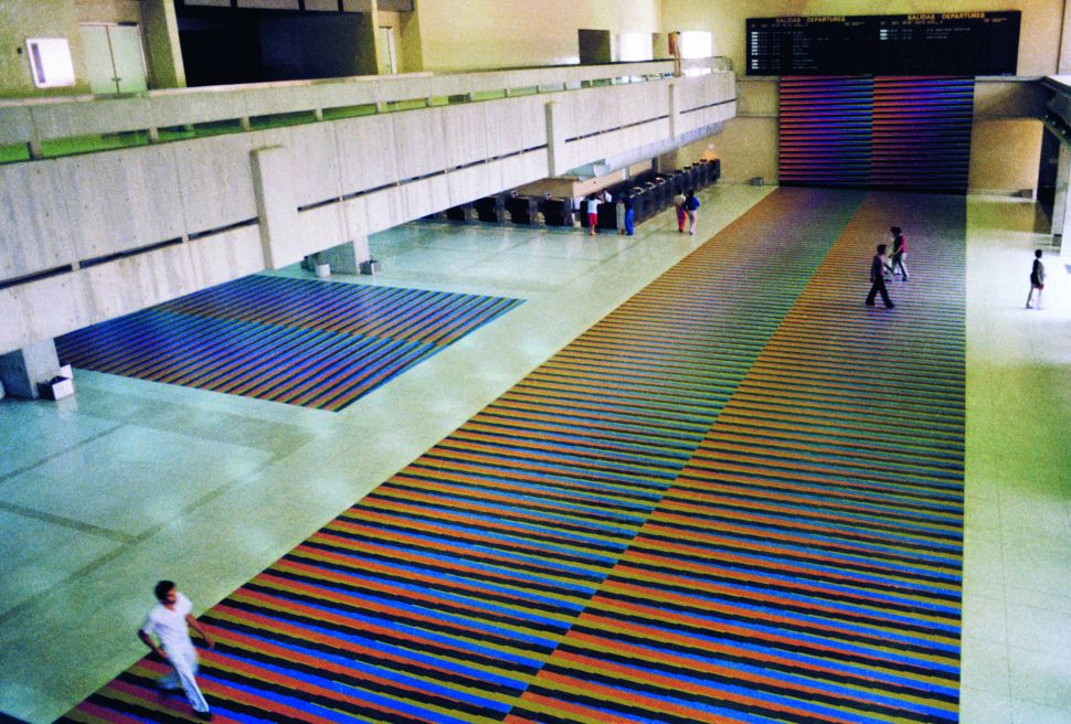 A vast airport hall with long bands of colored stripes covering the floor as travelers walk across the chromatic installation.