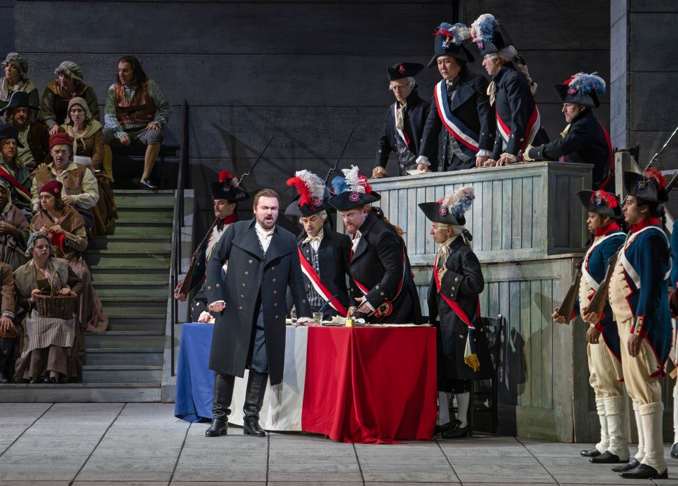 A scene from the Metropolitan Opera’s production of Andrea Chénier showing a crowded Revolutionary tribunal, with the baritone portraying Carlo Gérard standing at a tricolor-draped table as soldiers, officials, and onlookers surround him on multiple levels of the set.