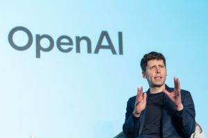 Man in black clothing sits onstage in front of blue screen reading 'OpenAI'