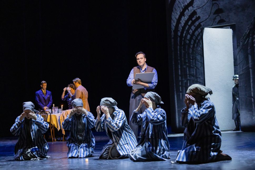 A dramatic moment from the opera shows several performers in striped concentration camp uniforms kneeling and covering their faces, while a man stands nearby holding a sketchpad and other actors converse at a dimly lit table in the background.