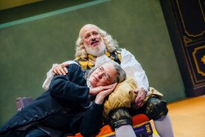 A bearded man in a gold-trimmed period costume embraces another man in dark clothing who rests his head sideways against the first man’s chest, both seated on a low bench in a stylized theatrical interior.