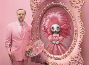Man in a pastel pink suit stands beside an ornate easel, holding a paint palette and brush. On the canvas is a surreal, wide-eyed doll with voluminous pink hair and a bow, framed in gilded pink. The studio background is entirely pink.