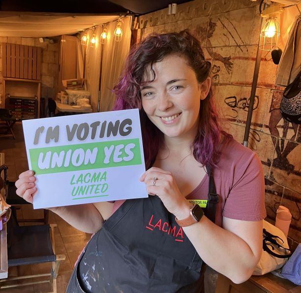 A museum educator wearing a LACMA apron smiles while holding a sign supporting unionization inside a workspace lit with warm lights.