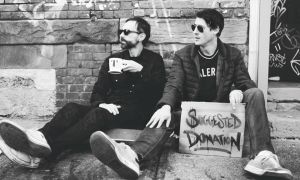 A black-and-white photograph of two men sitting against a graffiti-covered brick wall shows one drinking from a mug while the other holds a cardboard sign that reads "Suggested Donation," referencing the podcast of the same name.