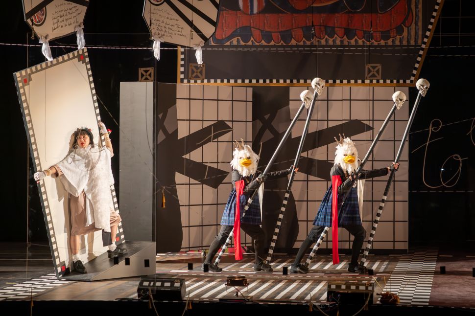 A group of performers wearing blindfolds and headpieces shaped like clustered spheres stand behind transparent panels with their hands raised, creating a choreographed tableau within a theatrical set.