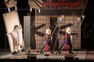 A group of performers wearing blindfolds and headpieces shaped like clustered spheres stand behind transparent panels with their hands raised, creating a choreographed tableau within a theatrical set.