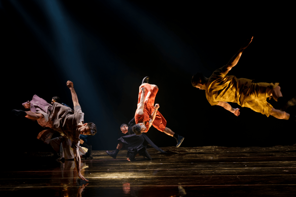 A group of dancers in earth-toned costumes leap and tumble across a dimly lit stage, their bodies captured midair above a reflective floor under focused beams of light.